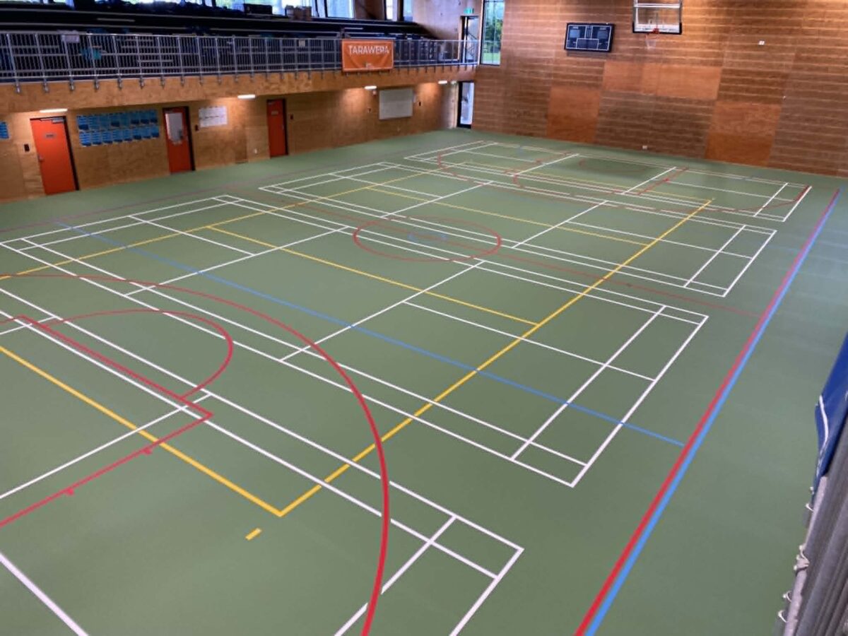 Olive green Herculan court at ACG Tauranga after re-topping, with dense white, red, yellow and blue multi-sport line marking, brick and timber walls and blue upper-level seating