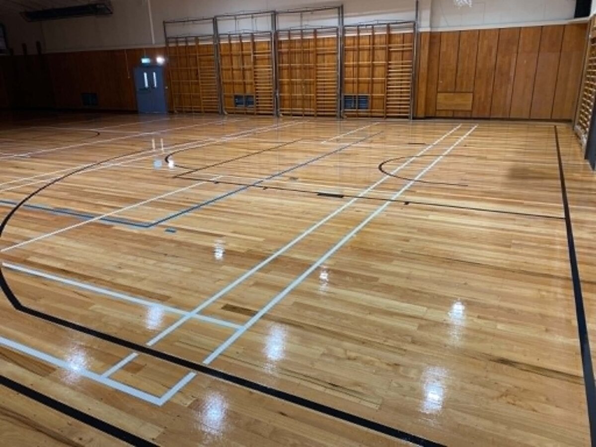 Freshly sanded pale maple timber gym floor at Cambridge College with a glossy finish and new line marking, timber wall bars along the back wall
