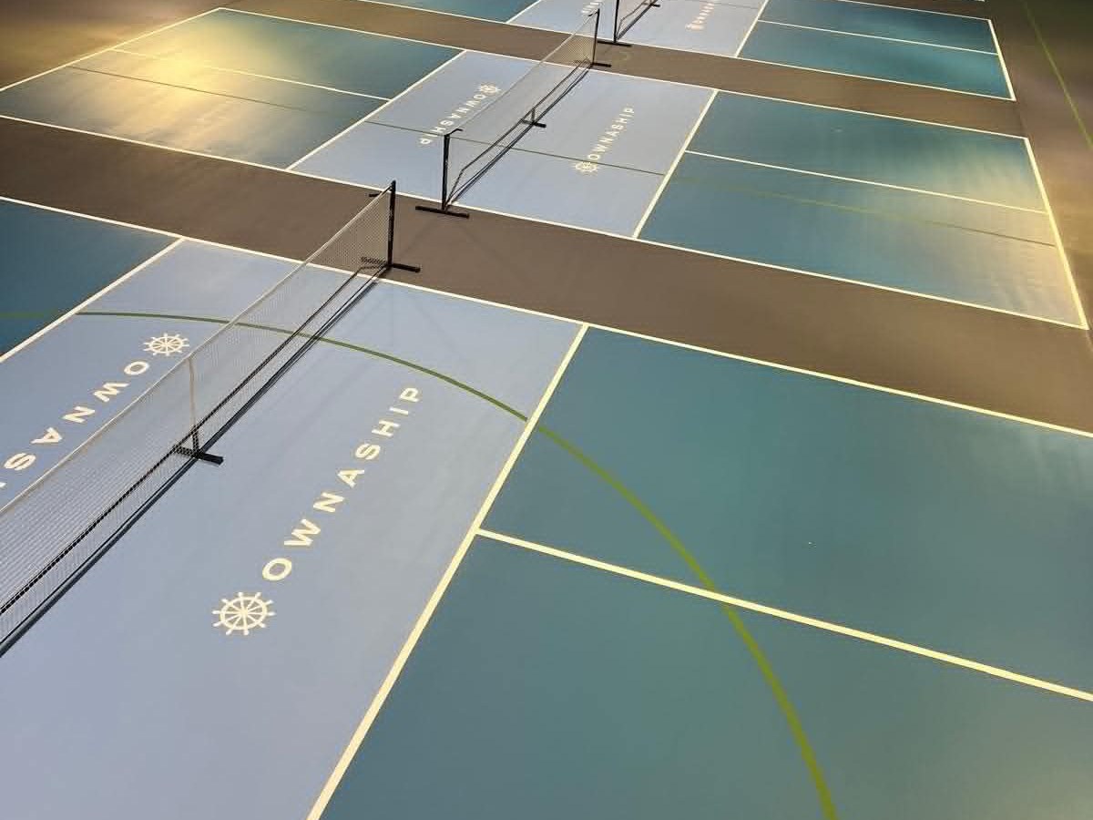 Several teal and grey pickleball courts at College Rifles with nets up and white line marking, under a white arched roof