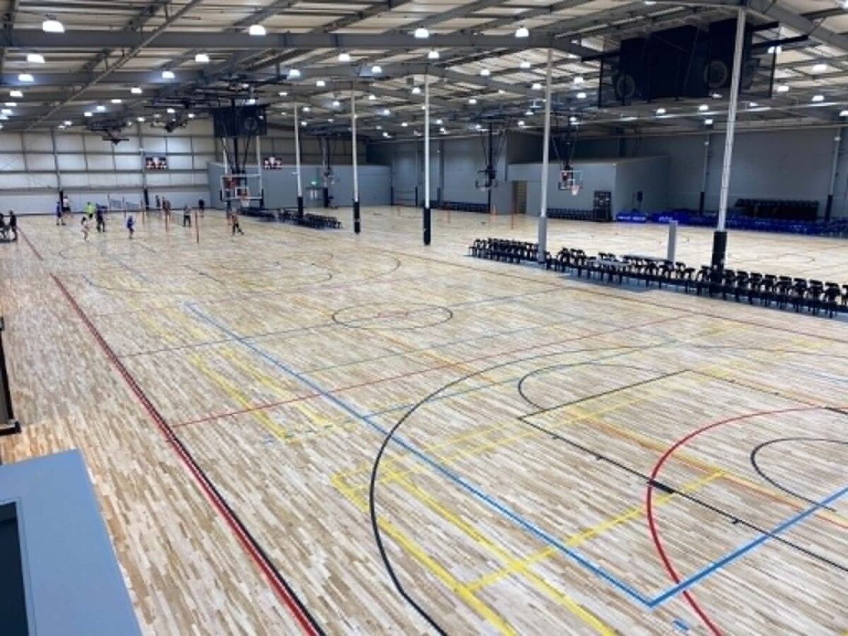 Large pale maple sprung timber court at Haumaru Sport & Recreation Centre in Tauranga with multi-colour line marking, players in the background and stacked seating under a high steel roof