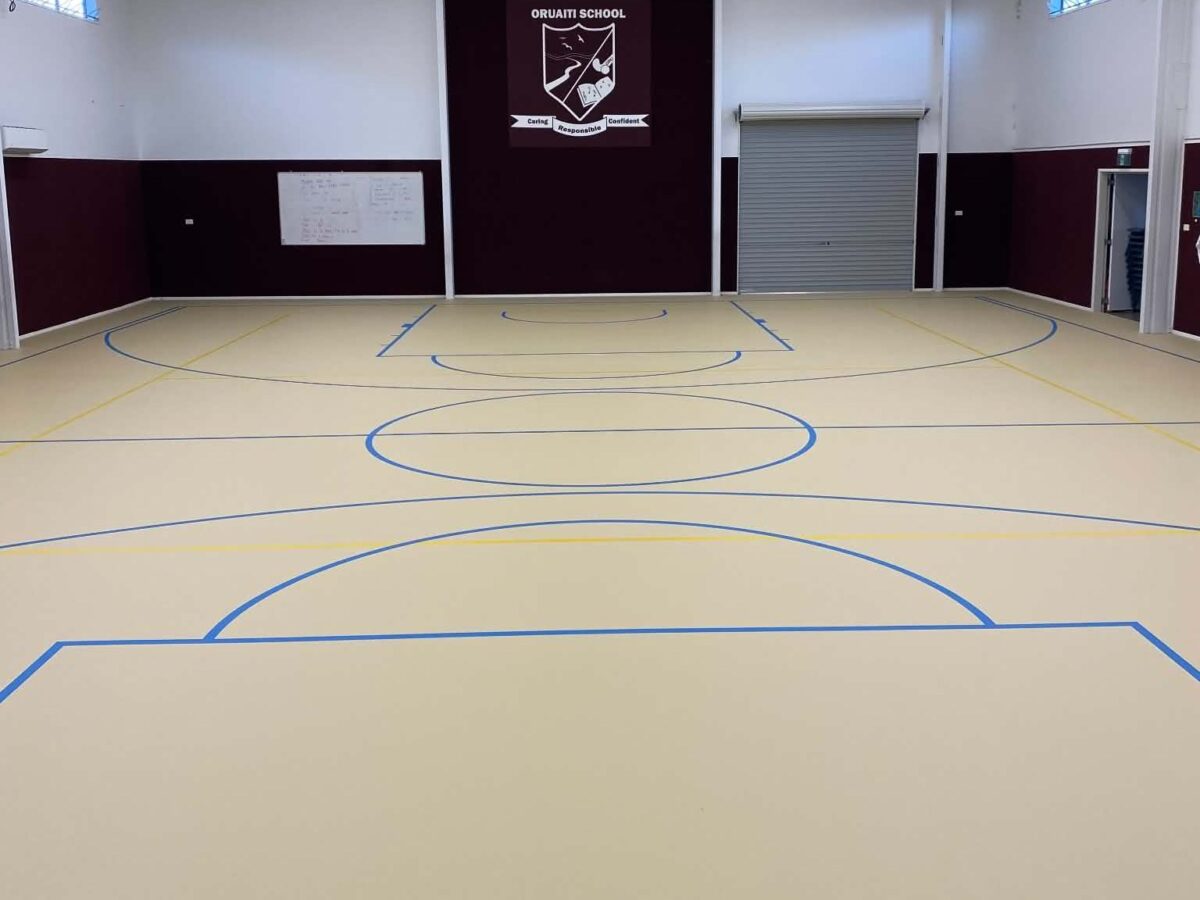 Cream Herculan floor with blue and yellow line marking in the Oruaiti School hall, maroon and white walls with the school crest on the end wall