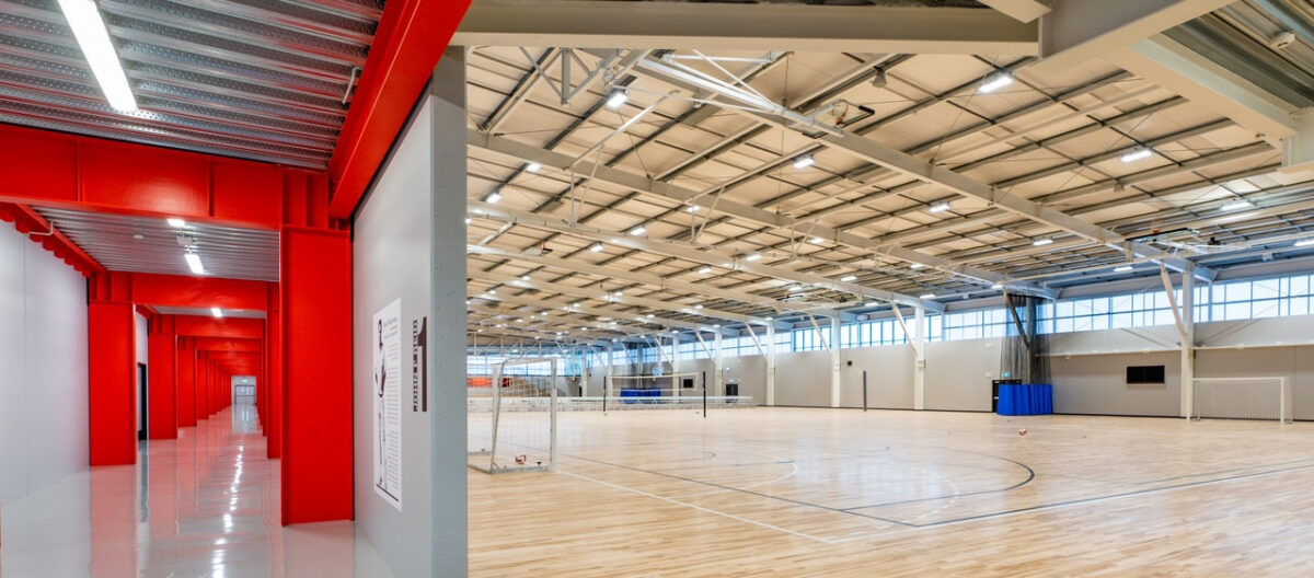 Maple sprung timber court at Selwyn Sports Centre seen past a red-painted corridor, with volleyball nets and high clerestory windows