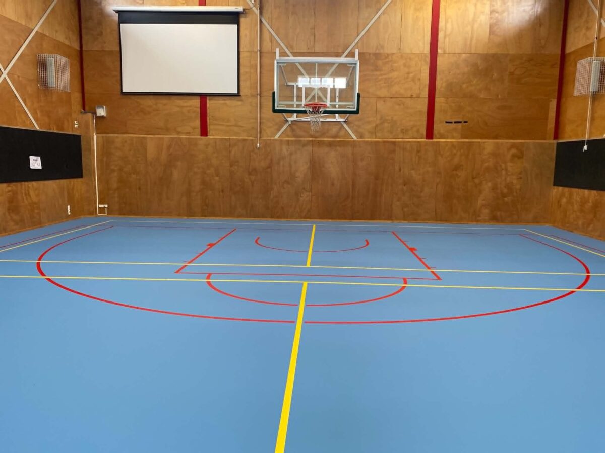 Light blue Herculan MF Blue 33 synthetic court at South Wellington Intermediate School with red and yellow line marking, plywood walls and a basketball backboard