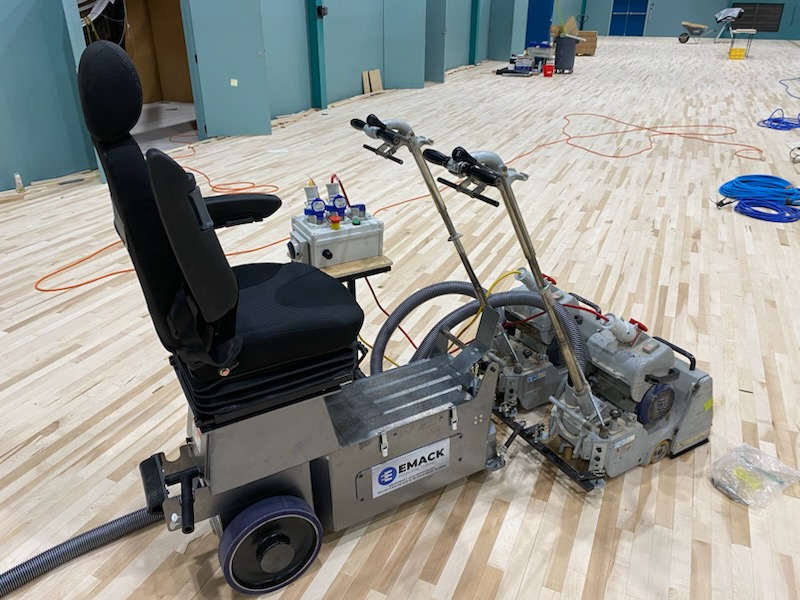 EMACK ride-on sanding machine parked on a maple sports floor part-way through a full sand, the timber stripped back to bare boards ready for re-coating