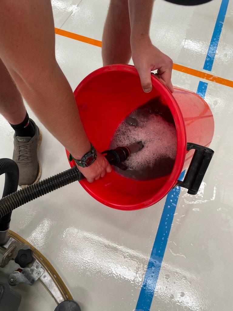 Technician emptying a red bucket of soapy water beside a scrubber hose on a freshly cleaned white sports floor, part-way through a scheduled clean