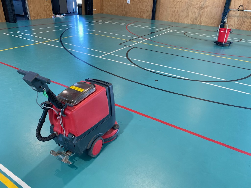 Red walk-behind scrubber part-way through a wet clean on a teal Herculan synthetic court, with a mop bucket standing by at the far end of the hall